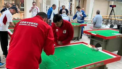 Phil Harrison/BBC Several men in different coloured tracksuits standing round waist-height tables which have green felt football pitches on them. The pitches have small goals and players.