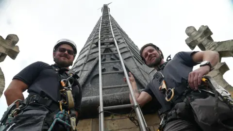 Joe Bilton/BBC Two men in black tops and shorts are wearing white helmets and harnesses. The photo is taken from the floor looking up. They are holding on to a ladder which ascends up a grey pinnacle with a weather vane at the top.