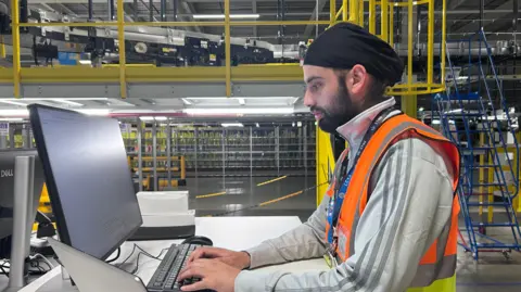Kate Bradbrook/BBC A man standing at a desk and typing at a computer. He is inside a warehouse and wearing a high visibility jacket