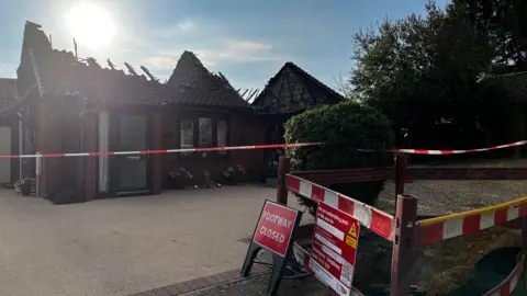 Jack Maclean/BBC A bungalow roof is destroyed by fire and cordons are put up around it, with a road sign saying that the path is closed