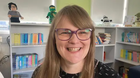 Miriam Donnan is wearing purple glasses, she has long brown hair. She is wearing a black shirt. Behind her are books on a shelf in a classroom. 