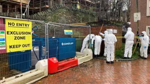 A group of police forensic investigators carrying poles, wearing white boiler suits. They are looking at a green bin. Next to them is temporary fencing with a yellow sign on it saying 'No entry, exclusion zone, keep out'