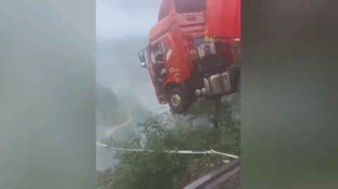 The cabin of a red lorry, with the driver leaning out of the open door, hangs over the edge of a broken bridge with a deep chasm below