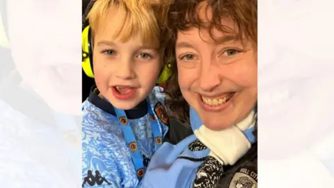 Submitted A young blond-haired boy smiles while taking a selfie with his mum, who has mid-length brown, curly hair. They both wear a blue coloured Hull City away kit and the boy wears fluorescent ear defenders. 