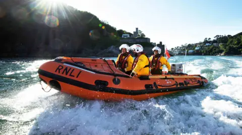 A lifeboat crew on the water on the river dart. The boat is orange with a black strip along the middle. The crew are all wearing yellow jackets and white helmets.