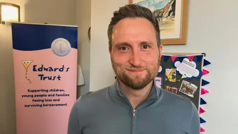 A man with dark hair and a grey beard is wearing a grey fleece in front of a blue and pink Edward's Trust sign near a wall.