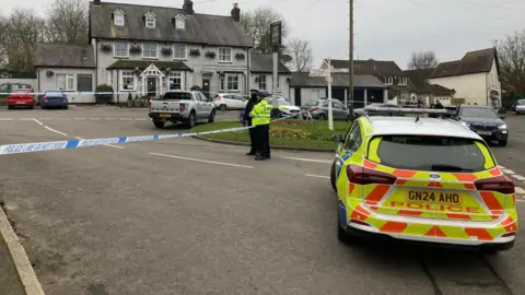 Dan MacLaren/BBC Police stand outside the pub, which is taped off. A police car is in the foreground