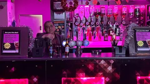 Kate Bradbrook/ BBC A bar area in purple ambient light with three red squares on the front. A man stands behind the bar. 