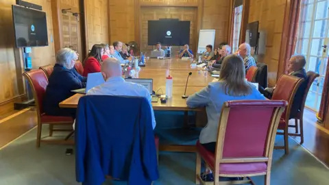 Matt Precey/BBC People sat around a large table in a committee room. The room has wood panels. There are three television screens positioned on the walls.