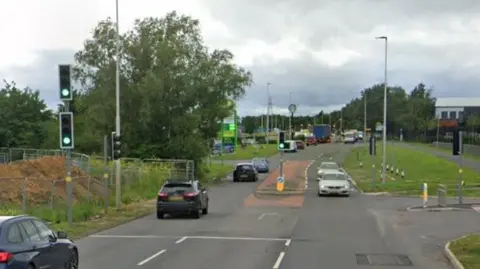 Cars driving in both directions through a traffic light junction on a busy main road through an industrial estate. A petrol station and a Starbucks can be seen in the distance.