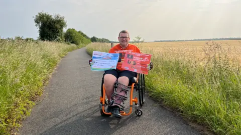 Tom Jackson/BBC A man in a wheelchair on the path where he started the challenge. The concrete path has overgrown grass on both sides. He is wearing glasses, has short brown hair and is smiling at the camera. His wheelchair has black wheels and an orange frame. His left leg is encased in an external cage.