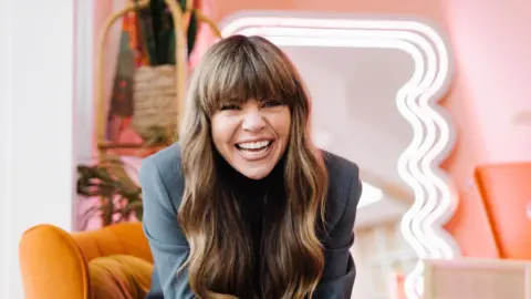 Briony May Williams smiling at the camera. She is wearing a grey suit jacket and has long brown hair which has loose waves. In the background is a pink and orange chair. 
