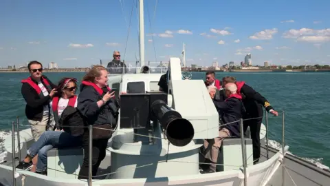 Looking at the boat from the bow, its large black gun barrel at the front. About eight people can be seen on the boat, including a couple of the veterans. Portsmouth harbour is in the distance.
