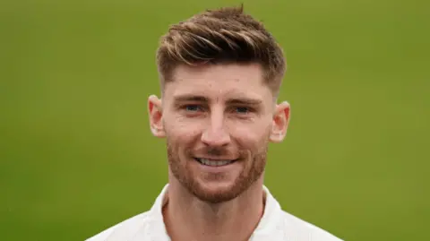 Rob Keogh wearing a white cricket shirt. He has light brown hair and stubble, and is smiling while standing on the field.