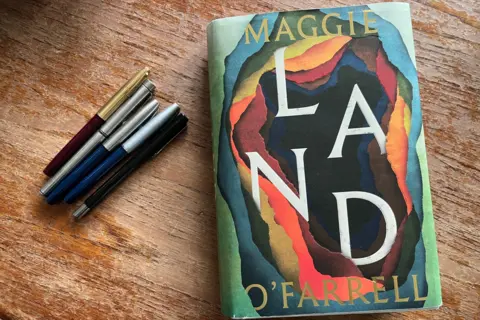 BBC/Katie Razzall 5 fountain pens on the left and copy of her new book Land on the right
front cover has bright colours of red, orange, blue and green