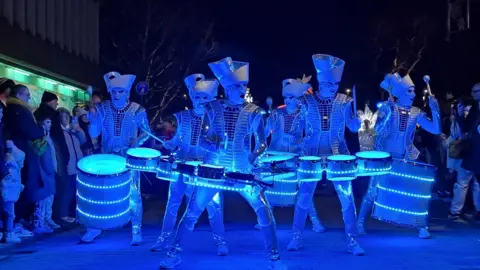 The Spark! LED drum band. They are drummers dressed in costumes that are lit up in blue LED lighting. Their steel drums are also lit up and wrapped in LED lights. They wear white face make-up and hats. They are walking down the street while playing music, with people lining the streets.