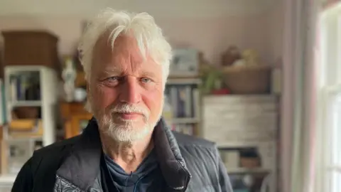 David Knopfler, wearing a black gilet, with a blurred background of shelves at his home