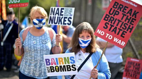 Getty Images Two women in Saltire facemasks hold signs saying "Bairns Not Bombs" "Nae Nukes" and "Break Up The British State No to War". One has blonde hair and the other dark. They are both wearing light blue tops with CND badges attached to them