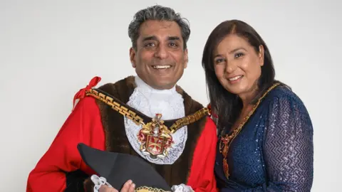 City of Wolverhampton Council The mayor is in the centre of the photo in mayoral clothing, which is partially red and white. His wife with long dark hair is on the right and wearing blue. They are both smiling at the camera.