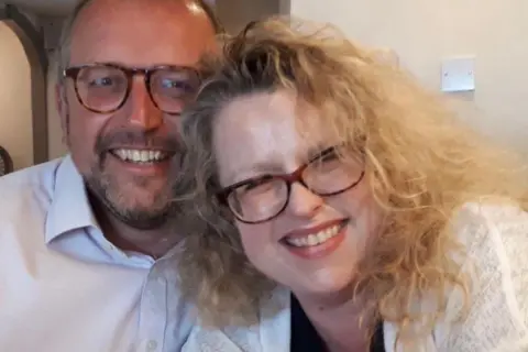 Richard Williman A man and a woman smile as they pose for a selfie. Both wear glasses and light-coloured tops. The man has short grey hair and a dark beard, the woman has curly blond hair and wears red lipstick.