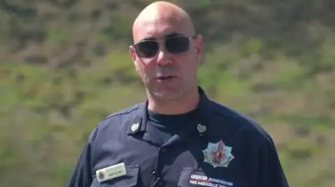 GMFRS Jim Willmott has shades, shaved head and a firefighter uniform. He stands on moorlands.