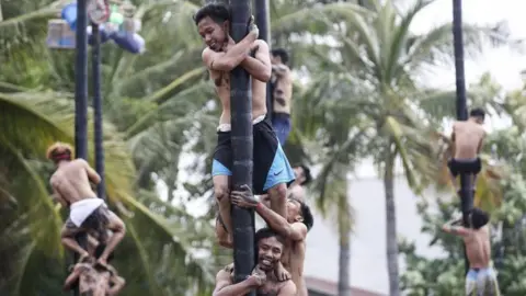 EPA Men climb greasy poles in a race. Some stand on each other's shoulders against a backdrop of palm trees. 