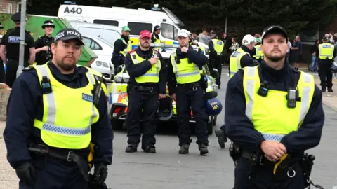 Uniformed police officers in high-visibility vests stand and walk along a residential street, with police vehicles parked nearby and members of the public gathered in the background.