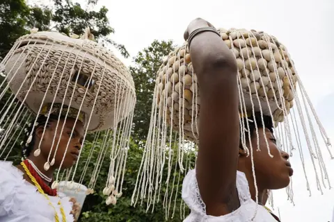 Anadolu via Getty Images Vestidas com trajes tradicionais, duas mulheres caminham uma atrás da outra no Bosque Sagrado em Osogbo, Nigéria, em 8 de agosto de 2025