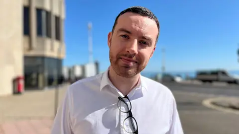George Carden/BBC Huw Ware stood outside the brighton centre with short brown hair and a white shirt.