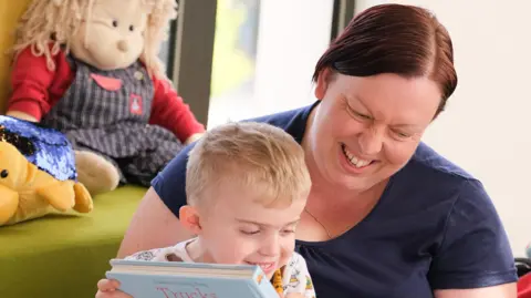 Anastasia Jobson/Sense Thomas is sitting on his mum's lap and is holding a book about trucks. They are both laughing. 