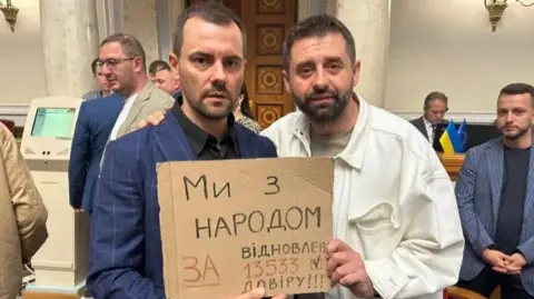 Dois homens, um de terno e outro em uma camisa branca, seguram uma placa de papelão no ucraniano