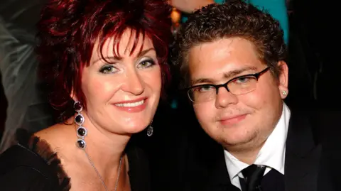 A close-up of Sharon Osbourne, left, with a younger Jack, right. Sharon is wearing a black low cut top and large dangling black earrings. Her hair is red and cut short and she is smiling. Jack is wearing a black suit and tie with white shirt, wire-framed glasses and diamond studs in his ears.