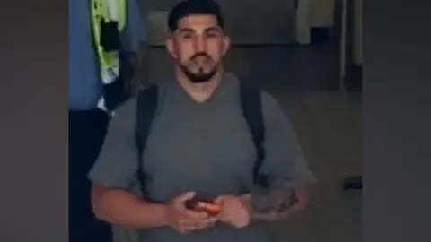 A man in a grey V-neck t-shrt hold a phone in both hands as he walks on a tiled floor in this CCTV still.