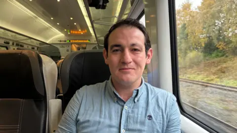 Nathan Mulholland sitting on the train. He has dark hair and wearing a light blue shirt and looking directly at the camera. The background of the train is on his right and the window of the train is on his left.