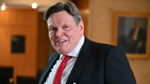 Getty Images Stephen Kerr, who has dark hair with a side parting, looks at the camera in a medium close-up shot in the Scottish Parliament. He is wearing a black suit, white shirt and red tie. 