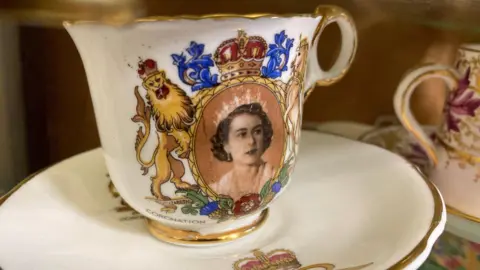 A ceramic coffee cup and matching saucer. The cup depicts Queen Elizabeth II as a young woman. The cup says "coronation" and has the royal coat of arms on it. The rim of the cup is gold.