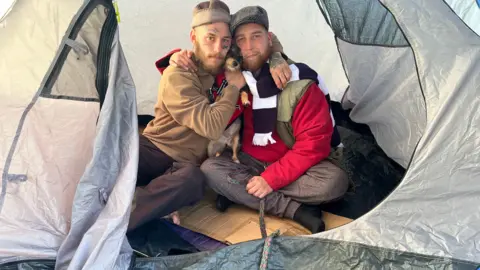 Two men with wearing hats are seen sitting inside a tent, cuddling a small dog.