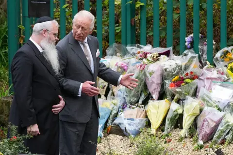Getty Images A man with a long white beard, white hair and glasses is with another man (King Charles III) - both are wearing suits. King Charles's hands are outstretched towards more than a dozen bunches of flowers laid on the ground against a fence.