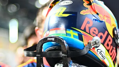 Getty Images The back of Arvid Lindblad's F1 helmet. It shows the three flags of England, white with a red cross, India's horizontal lines of orange, white and green with a blue circle in the middle then on the right, Sweden's blue background with a yellow cross. The helmet also features the Red Bull sponsor with a red bull and yellow background with the writing of the brand below it. You can see the top of Arvid's racing suit and a black support for protecting his neck in a crash. 