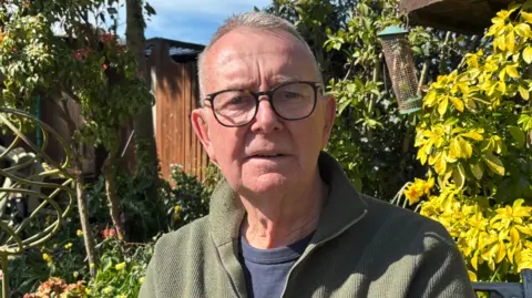 A man, with glasses and grey hair, in a green fleece with blue underneath is looking at the camera. Bushes are behind him.