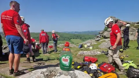 Devon Cave Rescue Organisation Rescue workers on a moor, with large granite boulders around, and a bottle of green washing up liquid on a rock in the foreground