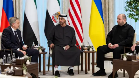 A handout photo released by the UAE Presidential Court shows UAE President Mohamed bin Zayed Al Nahyan (centre), meeting Rustem Umerov, Secretary of the National Security and Defense Council of Ukraine (R), Igor Kostyukov, Chief of the Main Directorate of the General Staff of the Russian Armed Forces (L)during trilateral talks between the United States, Russia and Ukraine at Al Shati Palace in Abu Dhabi. The participants are sitting in a semi-circle with the Russian, UAE, US and Ukrainian flags behind them.