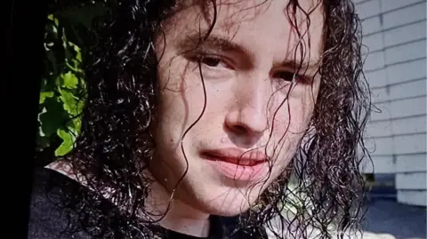 Vlad Nikolin-Caisley looks at the camera through his long, curly and black hair which hangs down to his shoulders. He is wearing a black t-shirt.