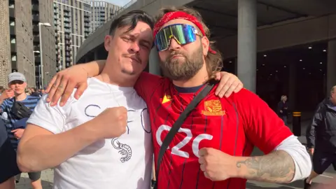 Henry Godfrey-Evans/BBC Owen Leworthy holding his fist up while wearing a white T-shirt. He has his arm round a man wearing a red Southend top and shiny sunglasses.