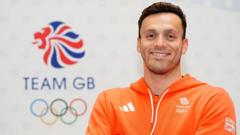 Image of James Guy MBE. He is pictured in front of an Olympic ring sign, with Team GB printed above it. He is wearing an orange hoodie with Team GB logos.