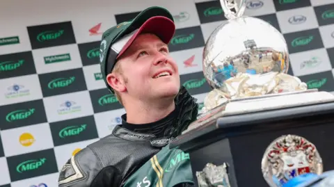 MANX GRAND PRIX Ingham holding the Senior trophy on the MGP podium. He is looking up and smiling.