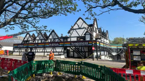 Kate Bradbrook/BBC A police officer is speaking to a man in high-vis clothing while a white building stands in the background with the visible roof.
