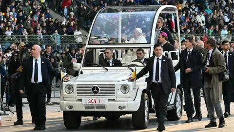 Getty Images The pope, waving to large crowds, sits in the white popemobile while many security people walk alongside the vehicle.