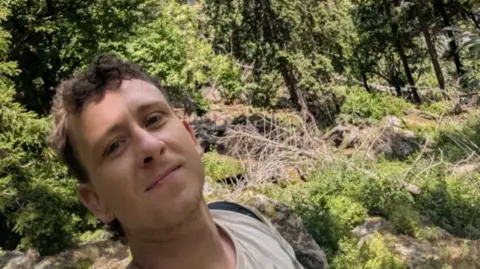 A man with short brown hair which is slightly curly on top taking a selfie surrounded by rocks, trees and general greenery. He is wearing a beige top and a black backpack.