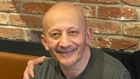 Jim Poptani Jim Poptani with very short hair and slight stubble, wearing a green T-shirt and smiling at the camera.  There is someone else's hand on his right shoulder, and he is sitting on a brown bench in front of a brick wall.